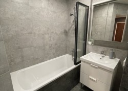 Paneláková kúpeľňa s vaňou a čierno rámovanou vaňovou zástenou, šedými obkladmi, bielou umývadlovou skrinkou a zrkadlovou skrinkou