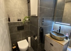 Paneláková toaleta v šedom obklade: závesné WC, nika na dekorácie a kamenné umývadlové umývadlo na drevenej doske; podsvietené zrkadlo