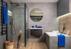 Veľká kúpeľňa s betónovým dekorom, vaňou obloženou šedými dlaždicami, závesnou skrinkou pod umývadlom a okrúhlym zrkadlom