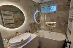 Malá kúpeľňa s oválnou vaňou, zlatými profilmi vaňovej zásteny, oválnym umývadlom na doske, podsvietenou nikou na kozmetiku a veľkým kruhovým zrkadlom