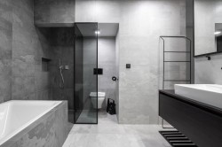 Čistá šedá kúpeľňa s obmurovanou vaňou, walk-in sprchovacím kútom, sklenenou priečkou k WC, tmavou umývadlovou skrinkou a veľkým zrkadlom cez stenu