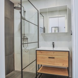 Sklenený sprchovací kút s čiernym rámom vedľa umývadlovej skrinky v drevodekore; svetlé obklady a zrkadlová skrinka nad umývadlom