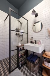Industriálna kúpeľňa s čierno rámovaným sprchovacím kútom, bielymi obkladmi, okrúhlym zrkadlom a úložným regálom s kozmetikou pri umývadle