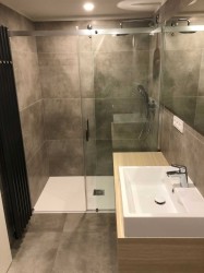 Menšia kúpeľňa so sprchovacím kútom, umývadlom na drevenej skrinke a tmavým rebríkovým radiátorom na šedom obklade