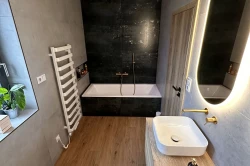 Menšia kúpeľňa so vstavanou vaňou v tmavom obklade, podlahou s drevodekorom, oválnym podsvieteným zrkadlom a zlatou batériou