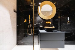 Čierna kúpeľňa so zlatými doplnkami, okrúhlym výrazným zrkadlom a umývadlom na skrinke; walk-in sprchovací kút.