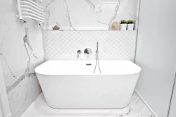 Voľne stojaca biela vaňa v úzkom priestore, mramorový dekor na stenách, jemná biela mozaika za vaňou a polička s rastlinami