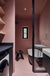 Minimalistická kúpeľňa v púdrovo ružovej s čiernou voľne stojacou vaňou, sklenenou zástenou, policami nas práčkou a malým oknom
