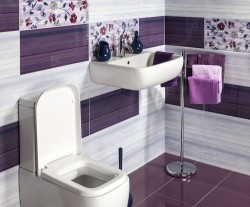 Závesné biele umývadlo a WC v kúpeľni s obkladmi v bielej a fialovej, doplnené kvetinovým dekorom a fialovými uterákmi