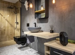 Detail modernej kúpeľne: kamenné umývadlo na svetlej drevenej doske, šedé steny, závesné čierne WC a sprcha s obkladom v dekore dreva