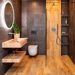 Menšia kúpeľňa s tmavými obkladmi, podsvieteným okrúhlym zrkadlom, závesným WC a sprchou s výrazným dreveným panelom a vstavanými policami