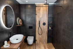 Tmavá kúpeľňa s veľkými obkladmi, oválnym umývadlom na drevenej doske, čiernou sprchovou batériou a sprchovacím kútom