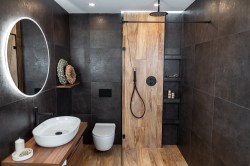 Tmavá kúpeľňa s oválnym podsvieteným zrkadlom, bielym umývadlom na drevenej doske a sprchou v drevodekore s čiernymi prvkami