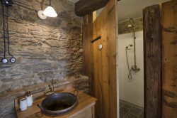 Kamenná stena a masívne drevené dvere pri sprchovacom kúte, okrúhle umývadlo na drevenej skrinke a mosadzné batérie vo vidieckom štýle
